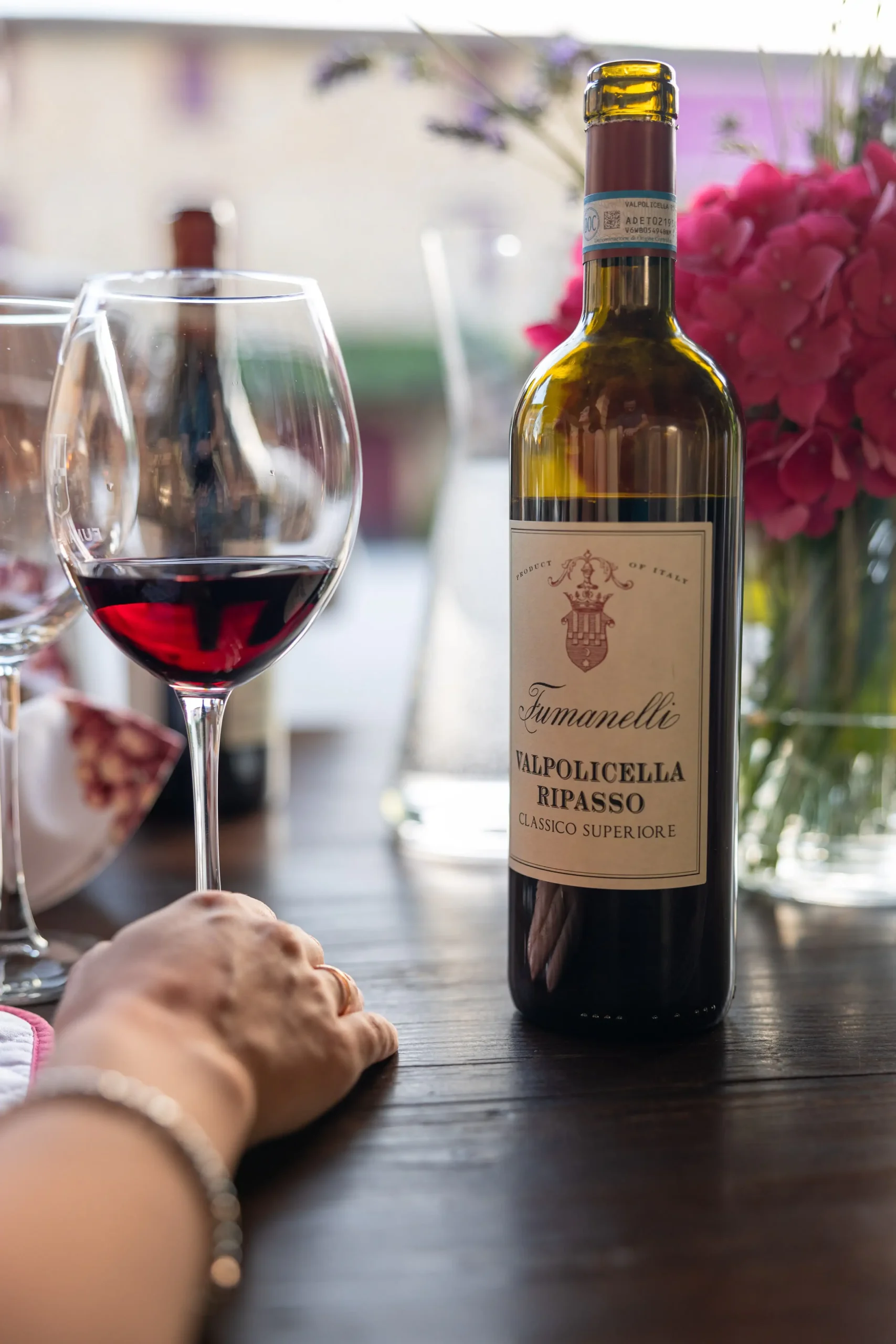 Bottiglia di Valpolicella Ripasso Classico Superiore Fumanelli su tavolo con calice di vino rosso e mano femminile in primo piano.