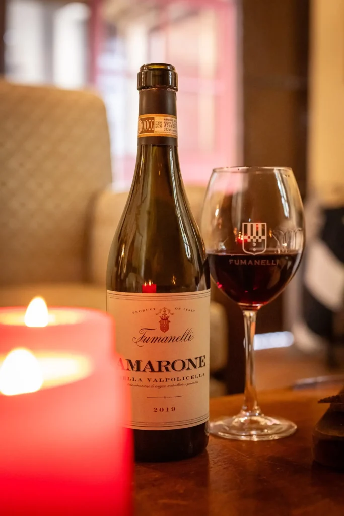 Bottiglia di Amarone della Valpolicella Fumanelli 2019 con calice di vino rosso e candela accesa in primo piano.