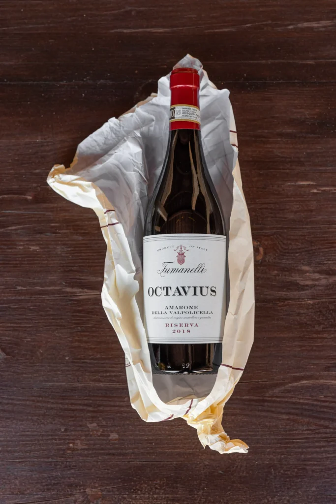 Bottiglia di Amarone della Valpolicella Riserva Octavius Fumanelli 2018 avvolta in carta, su tavolo in legno scuro.