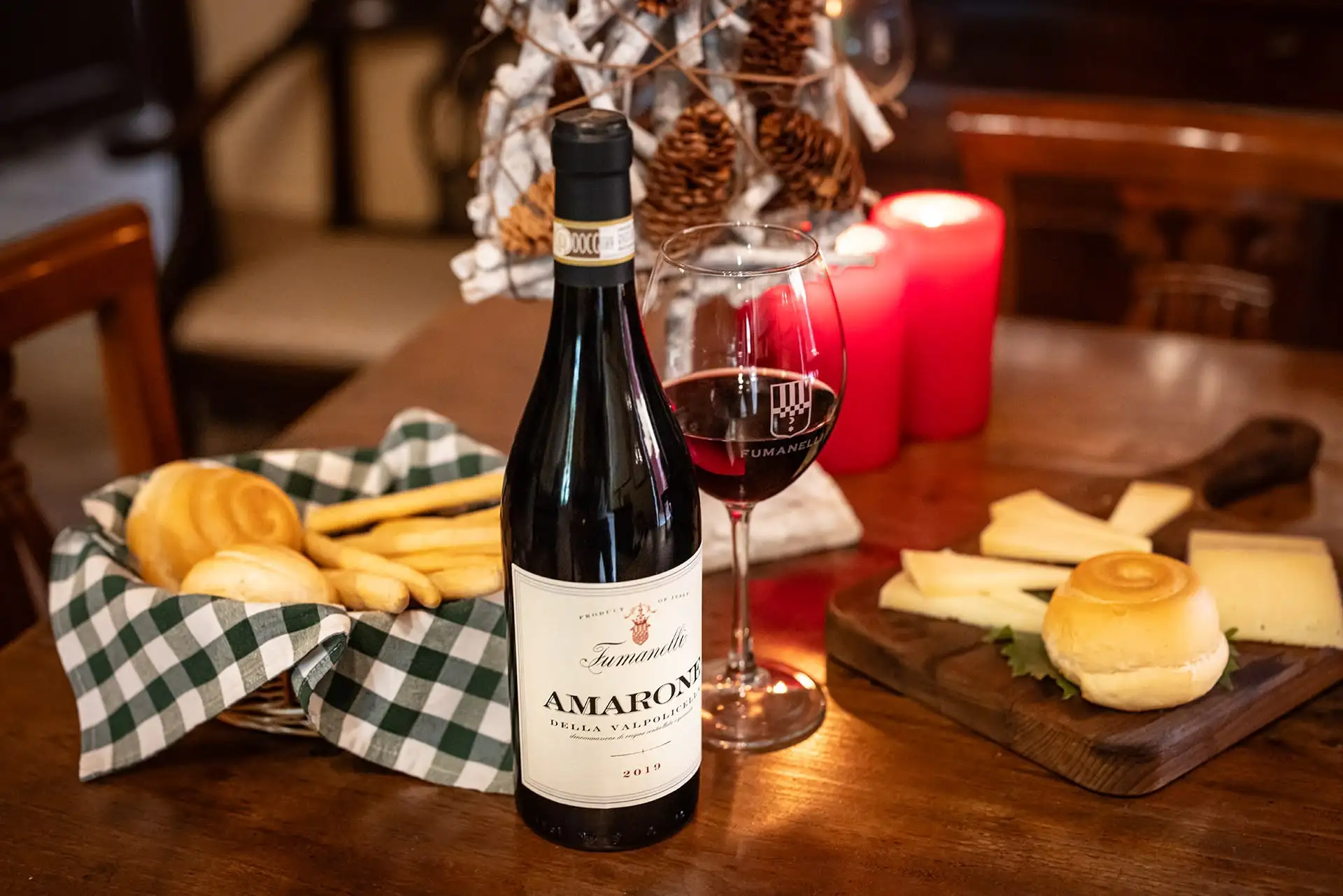 Bottiglia di Amarone della Valpolicella Fumanelli 2019 con calice di vino rosso, pane, grissini e formaggi su tavolo in legno.