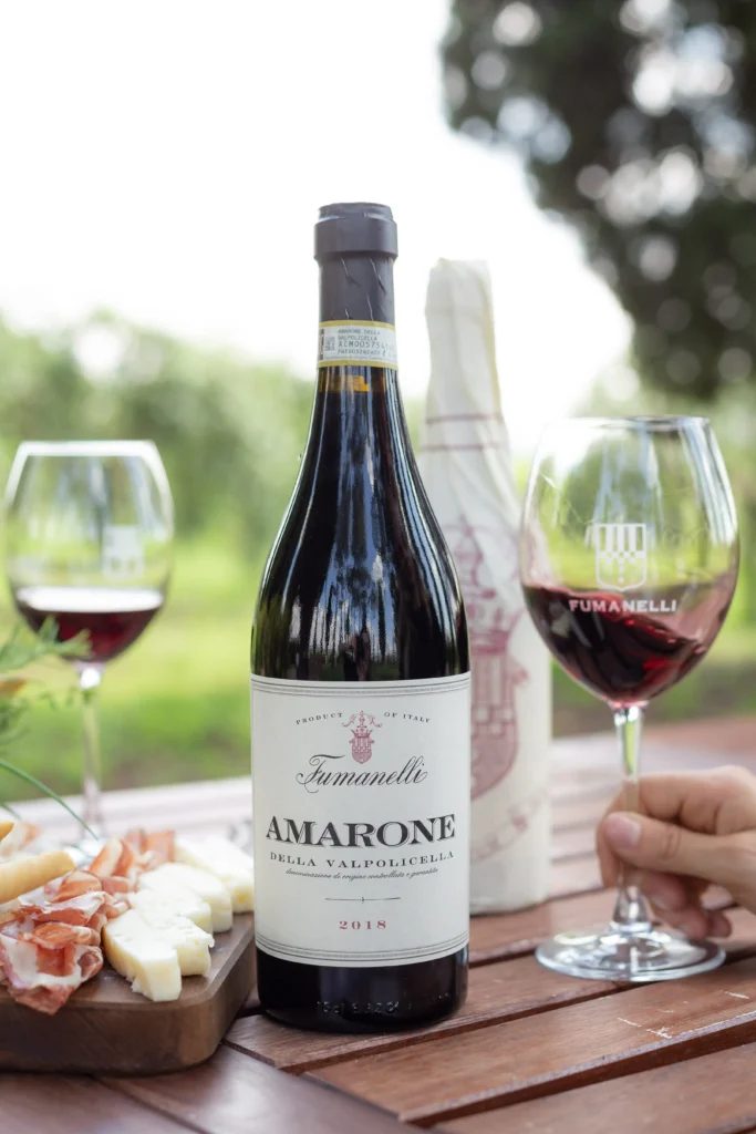 Bottiglia di Amarone della Valpolicella Fumanelli 2018 con calici di vino rosso e tagliere di salumi e formaggi su tavolo all’aperto.