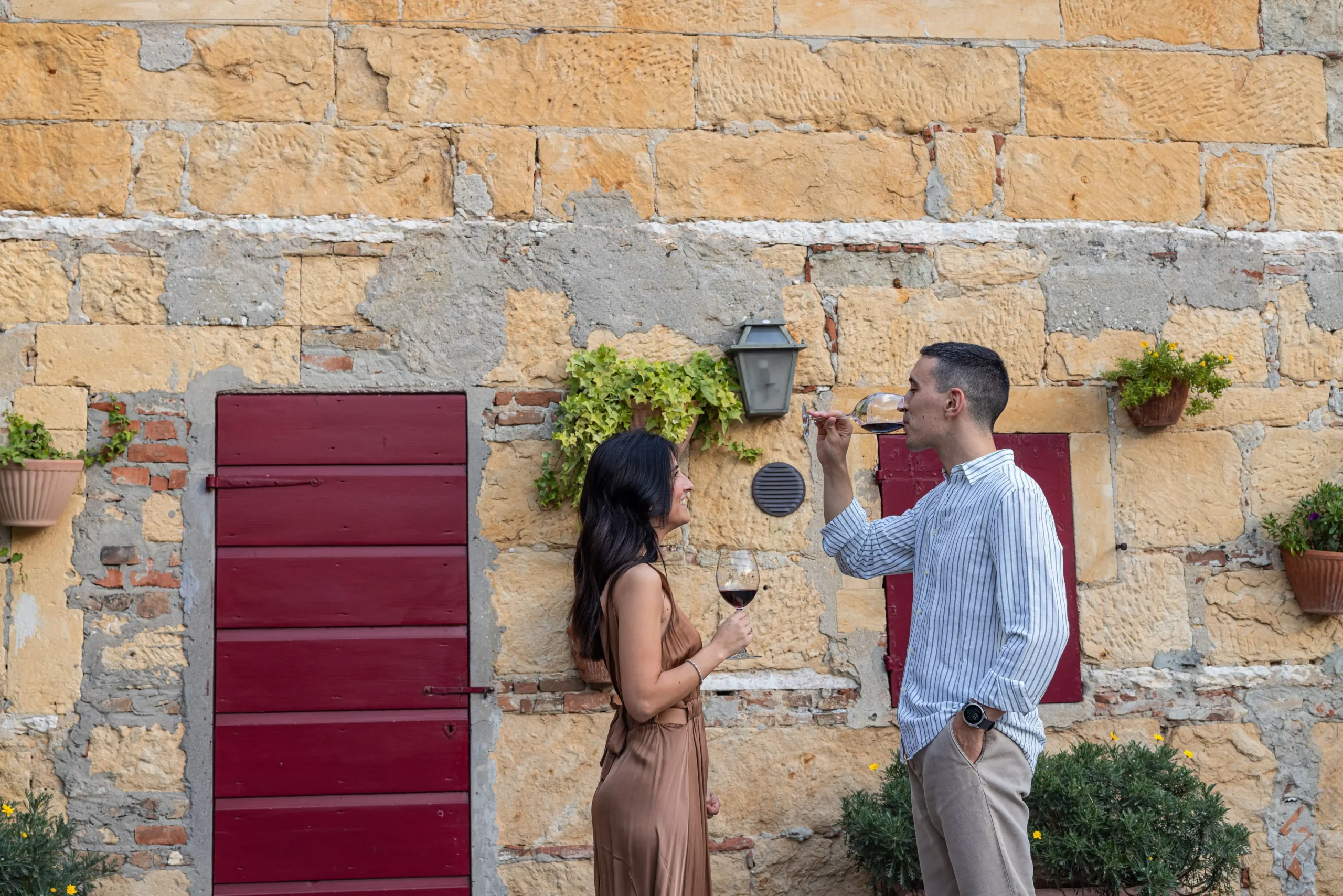 Coppia che brinda con calici di vino rosso Fumanelli davanti a una parete rustica con porte e finestre rosse.