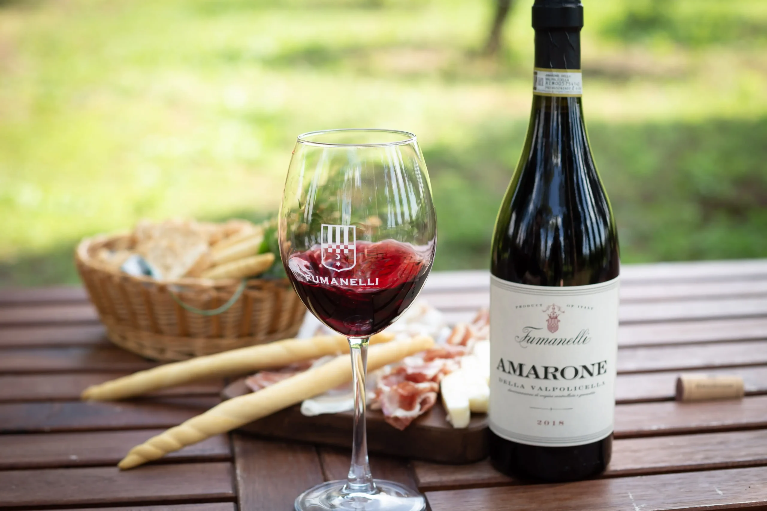 Calice di vino rosso Fumanelli e bottiglia di Amarone della Valpolicella 2018 con grissini, salumi e formaggi su tavolo all’aperto.