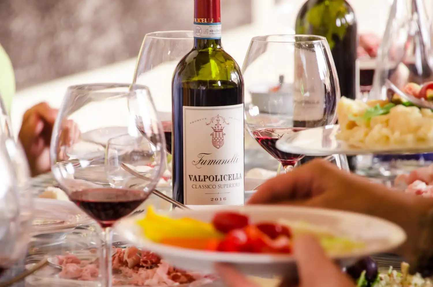 Bottiglia di Valpolicella Classico Superiore Fumanelli 2016 con calici di vino rosso e piatti di salumi e verdure su tavola conviviale.